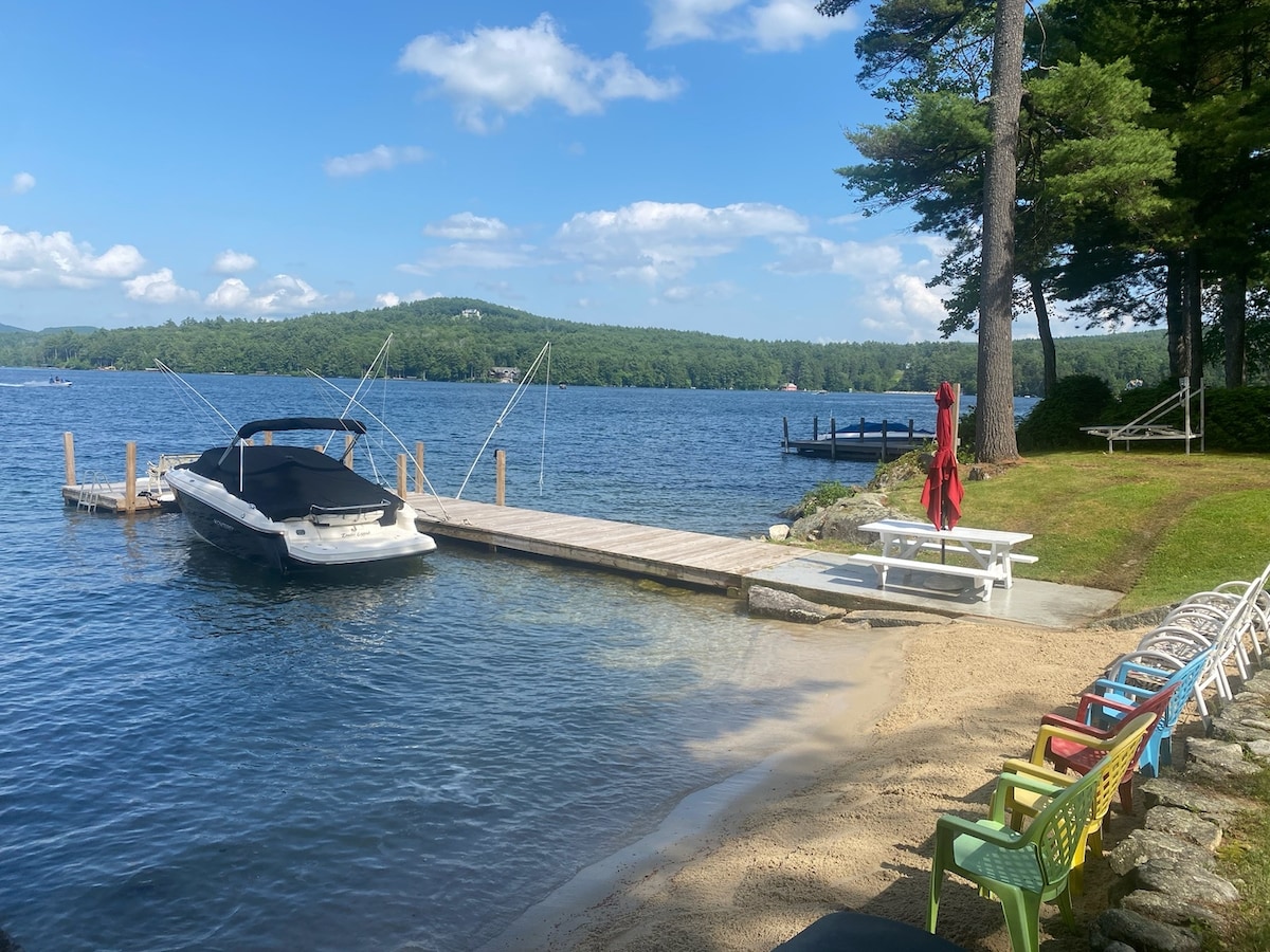 A serene lakeside view showcases a private sandy beach and a dock with a boat secured alongside. Colorful chairs are arranged neatly along the shore, with lush green trees framing the tranquil waters and distant hills under a clear blue sky.