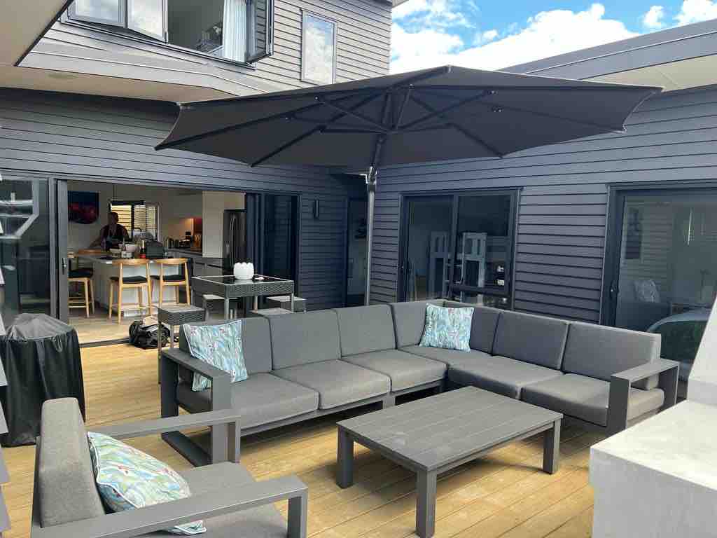 A spacious outdoor living area features a large sectional couch with cushions, arranged around a low table. A large umbrella provides shade, while the enclosed space offers accessibility to the interior. The surroundings include wooden decking and large glass doors leading inside.