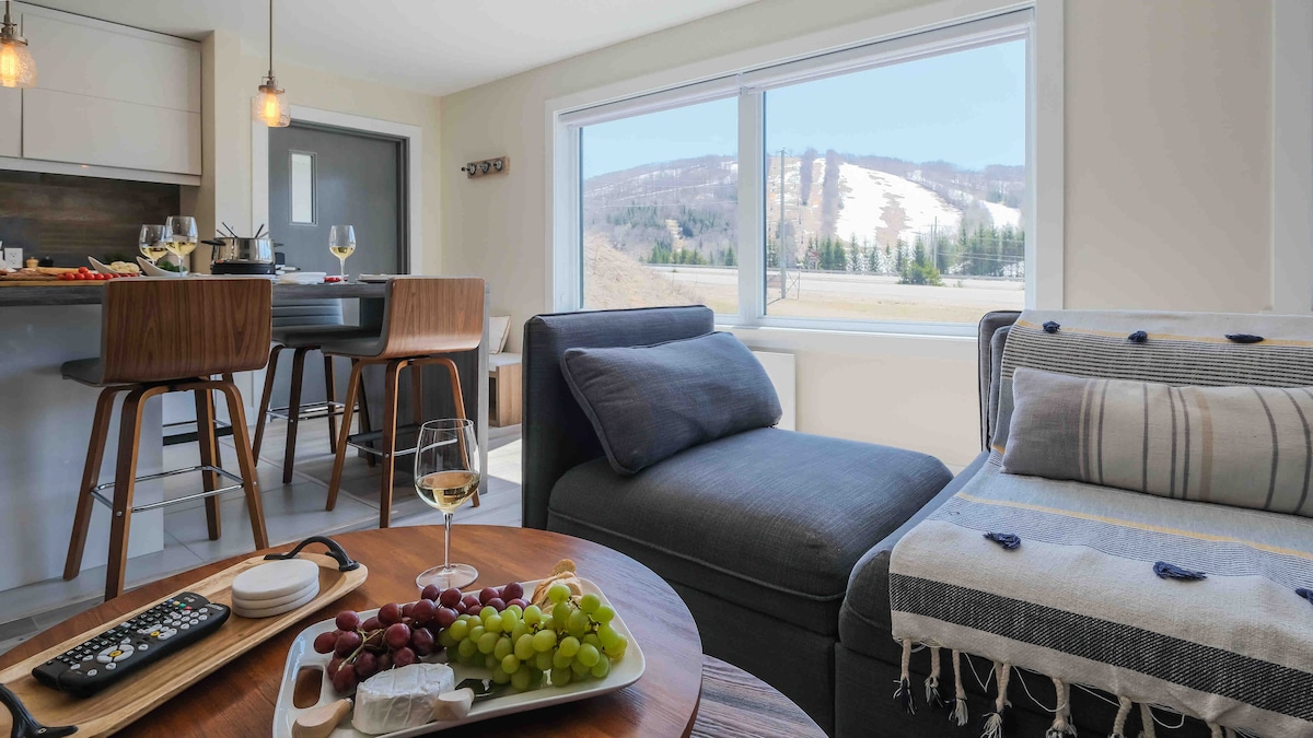 The living space features a comfortable seating area with soft furnishings. A table is set with a platter of fruit and cheese, while large windows provide a view of the nearby hillside. Light pours in, creating a bright and airy atmosphere.