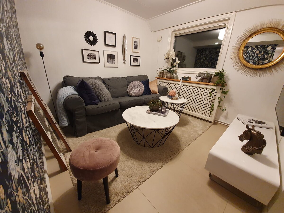 A cozy lounge area features a three-seated sofa adorned with various cushions. Two marble coffee tables are positioned on a soft rug, with plants and decorative accents enhancing the space. A large window allows natural light, and wall art adds a personal touch.