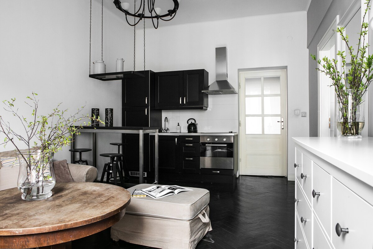 An open kitchen and living area features dark cabinetry and a modern stainless-steel stove. A round wooden table is complemented by a neutral-toned sofa. Small stools are positioned near a breakfast bar, while natural light enters through a window next to the door.