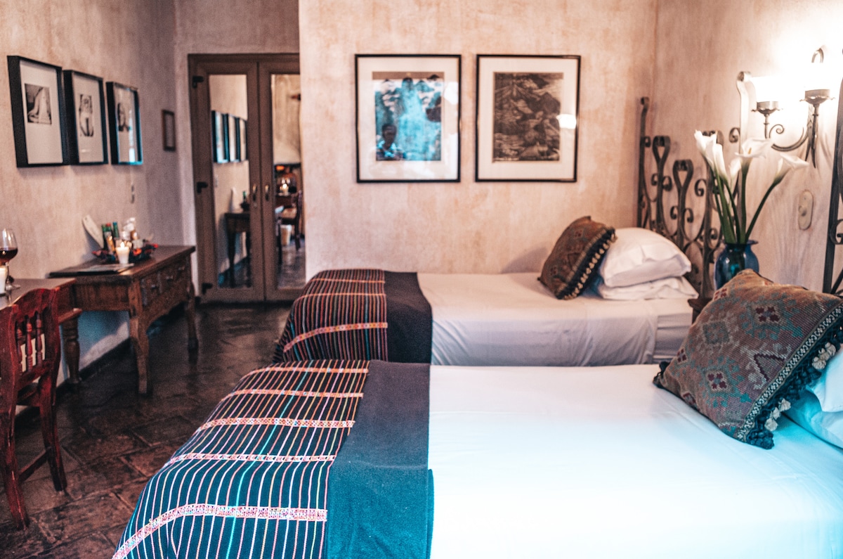 A tranquil room features two twin size beds framed by wrought iron headboards, adorned with woven blankets. A rustic writing desk rests against the wall, complemented by framed artwork. Soft lighting creates a warm ambiance, inviting relaxation and comfort.