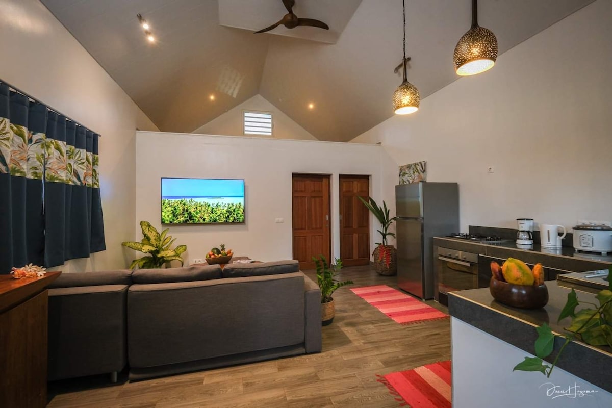 An open living area features a spacious grey sofa facing a mounted TV, with tropical-themed curtains framing large windows. Kitchen appliances and dining essentials are readily accessible, complemented by decorative plants and softly lit pendant lights.