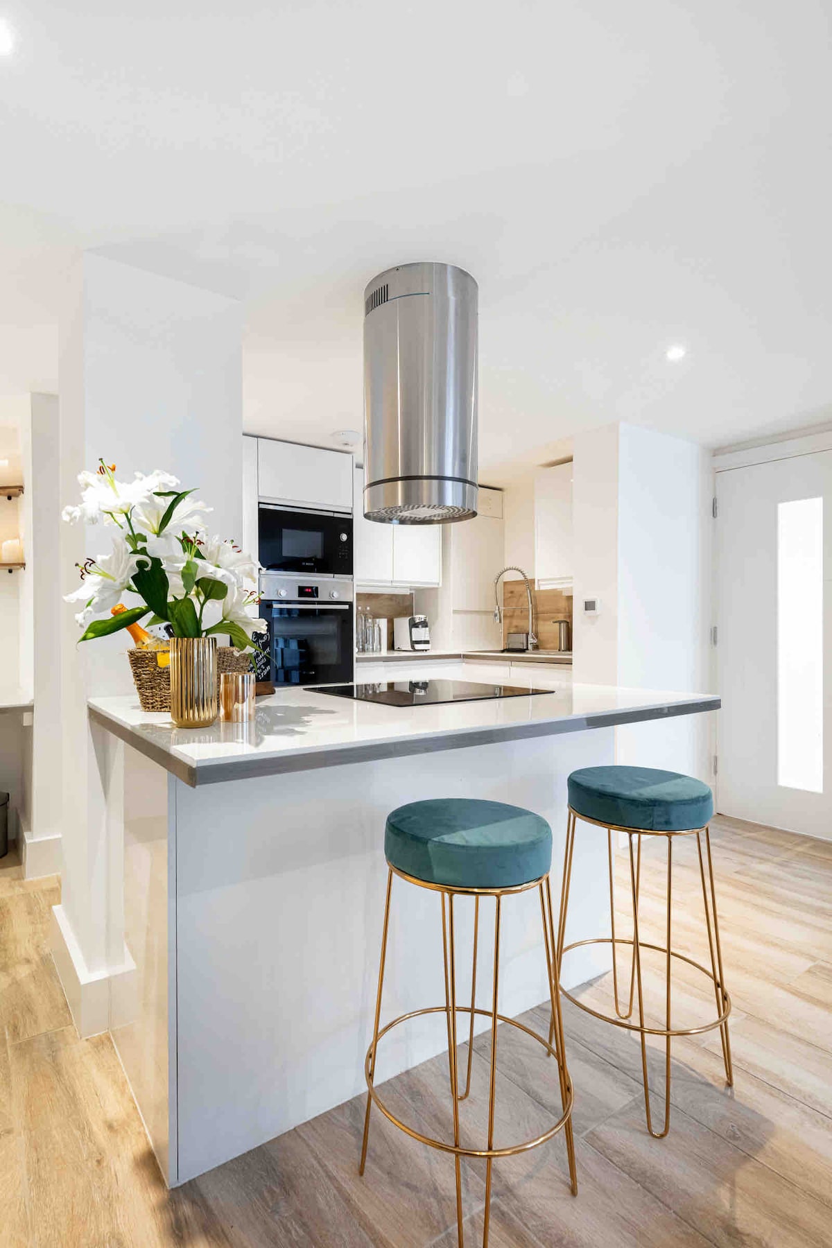 An open-plan kitchen area is showcased, featuring modern appliances integrated into sleek cabinetry. A stylish island bar with two teal velvet stools provides seating. A decorative arrangement of orchids adds a touch of nature, while a stainless steel hood adds a polished finish above the cooktop.