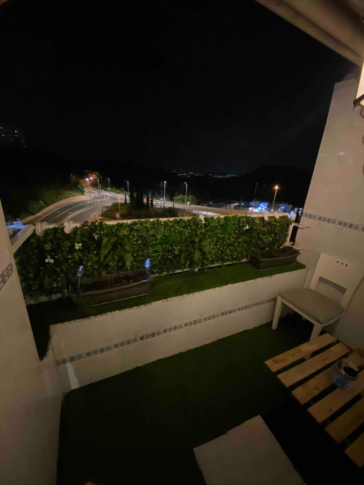 A balcony scene is presented at night, featuring green foliage along the walls. A small wooden table and a chair are visible, set against a backdrop of softly illuminated streets and distant hills.