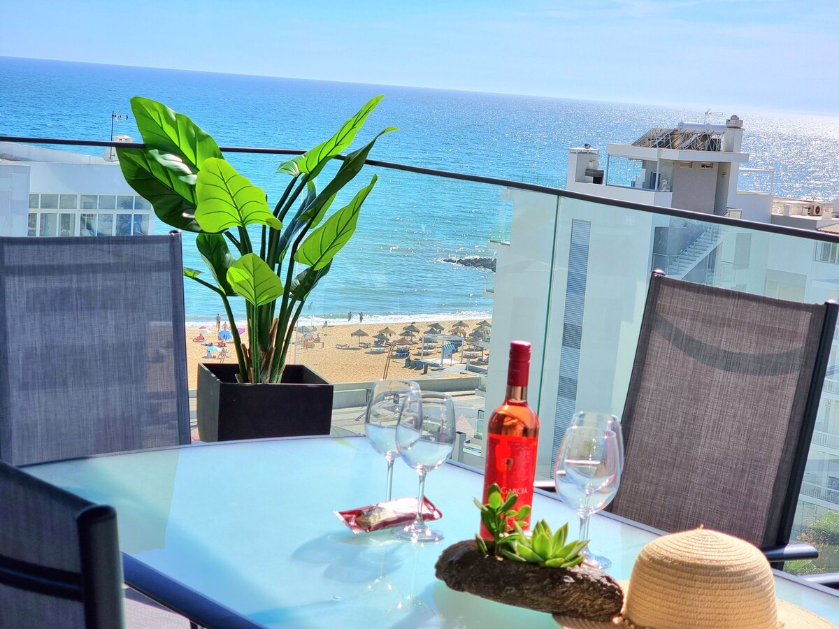 A terrace is showcased with a glass table and four chairs, overlooking the ocean and sandy beach. A potted plant adds greenery, while a bottle of wine and glasses are set on the table, enhancing the inviting atmosphere with distant umbrellas visible along the shoreline.