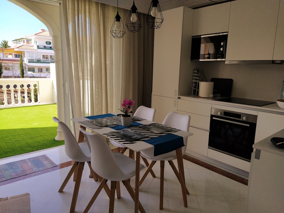A modern dining area features a white table with four chairs, complemented by a blue table runner and a small vase with pink flowers. Light floods in through large glass doors, leading to a grassy outdoor space surrounded by greenery.
