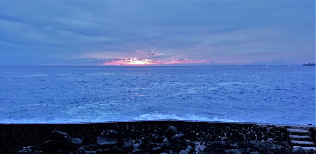 A serene ocean view is captured at sunset, with soft colors reflecting on the water. The rocky seawall is visible at the foreground, while the horizon displays a gradient of blues and purples, indicating the transition from day to night.