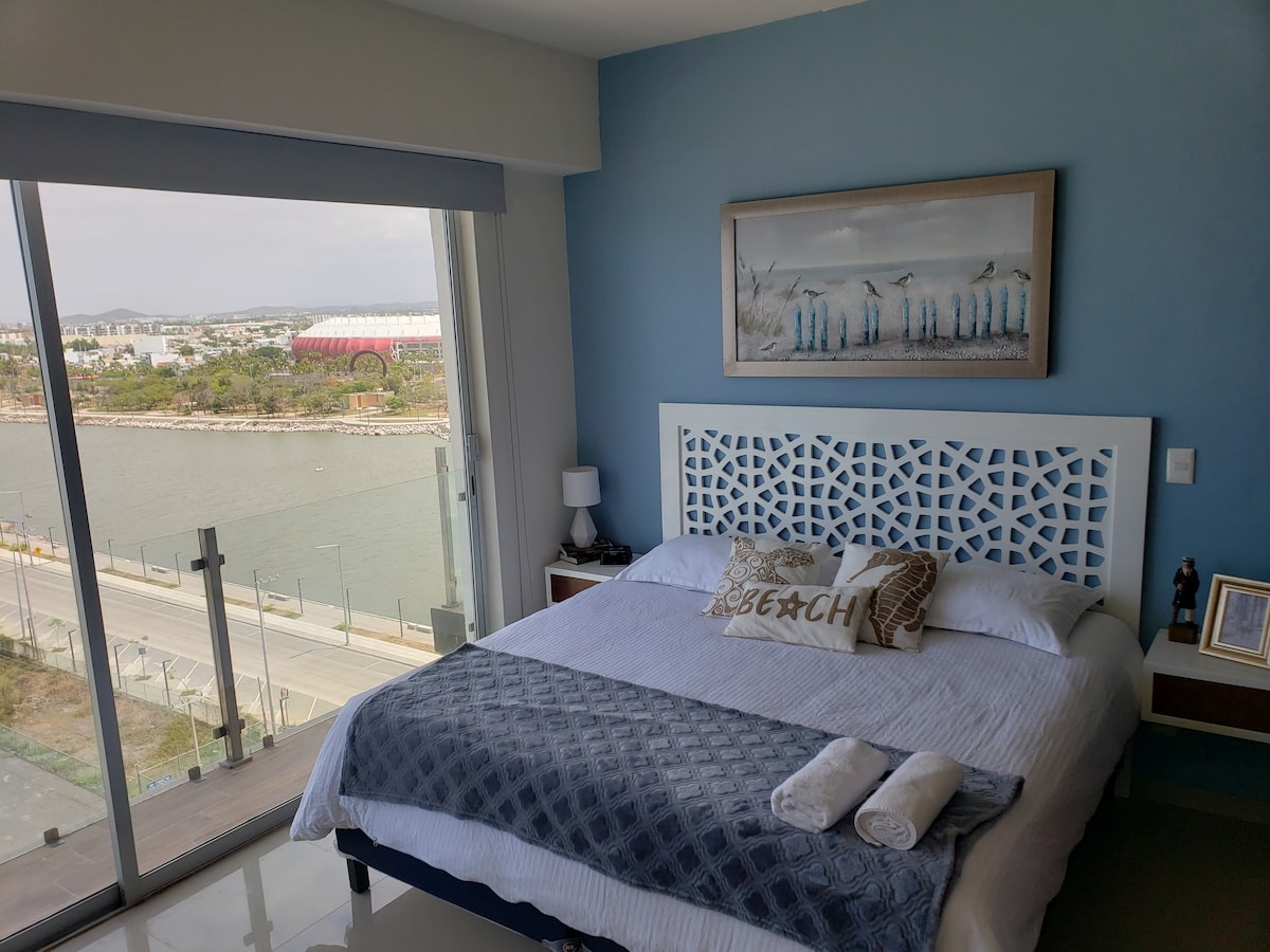 The bedroom features a large bed with decorative pillows and a cozy blanket, complemented by a nearby lamp and framed artwork. A glass wall offers scenic views of the waterfront, enhancing the natural light that fills the space.
