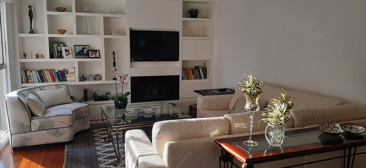 A living area features a comfortable sofa arrangement with a glass coffee table at the center. A television is mounted on a white wall unit, surrounded by shelves displaying decorative items. Natural light fills the space, accentuating the warm wood flooring.