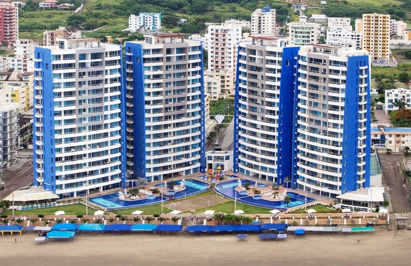 Four modern buildings with a striking blue facade are arranged around a shared outdoor space featuring two swimming pools and lounge areas. Beach cabanas line the sandy shore, providing shaded seating. The beach is just steps away from the complex, enhancing accessibility to waterfront views.