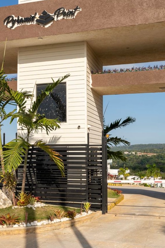 A modern entrance gate to the Pyramid Point community is framed by tropical plants and decorative landscaping. The structure features a combination of wood and metal elements, with greenery peeking from overhead, set against a clear blue sky in the background.