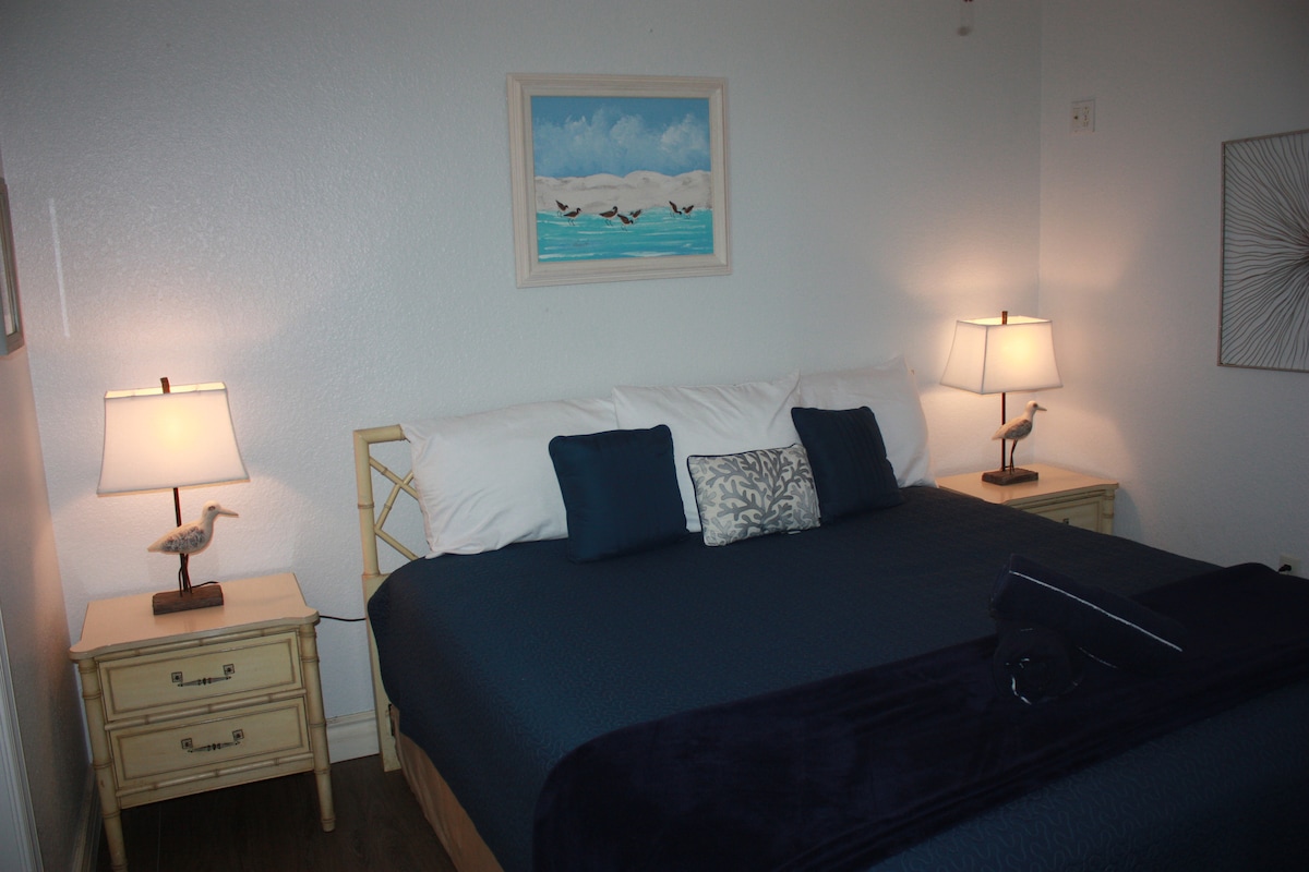 A spacious bed is positioned in the center of the room, adorned with white and dark blue linens. Two bedside tables are on either side, each featuring a lamp. A framed ocean scene hangs above the bed, complementing the serene color palette.