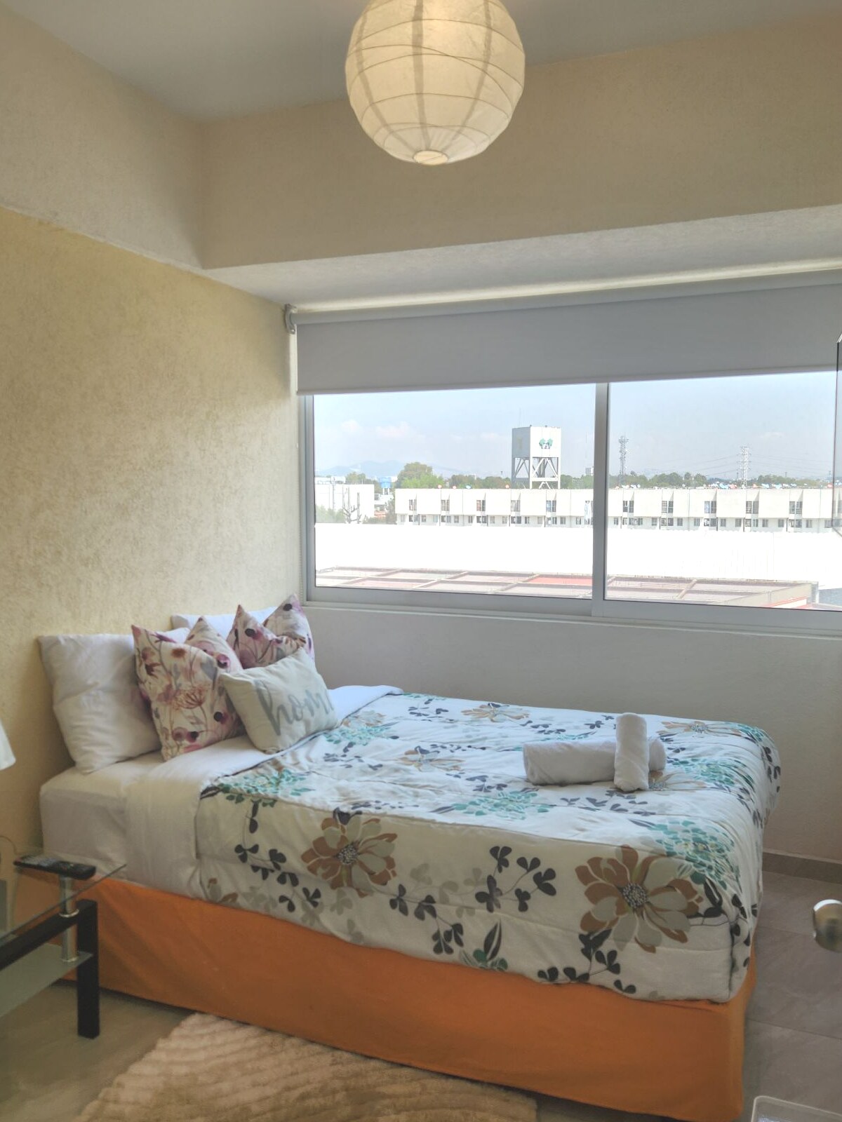 A bright sleeping area features a spacious bed adorned with floral bedding and accent pillows. Natural light streams through a large window, complementing the warm yellow walls. A simple bedside table and modern light fixture enhance the space.