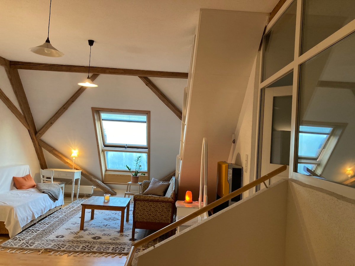 An airy loft space is highlighted by wooden beam accents and large windows. A cozy sitting area features a light-colored sofa and a patterned rug. Natural light floods in, reflecting off a nearby mirror. Stairs lead to a separate area, enhancing the spacious layout.