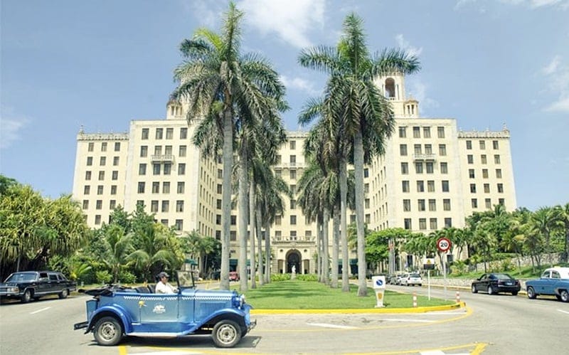 A grand hotel building is shown, featuring a classic architectural style with multiple floors and a prominent entrance. Palm trees line the pathway leading to the building, creating a lush green environment. A vintage car is seen driving on the street in front of the hotel.
