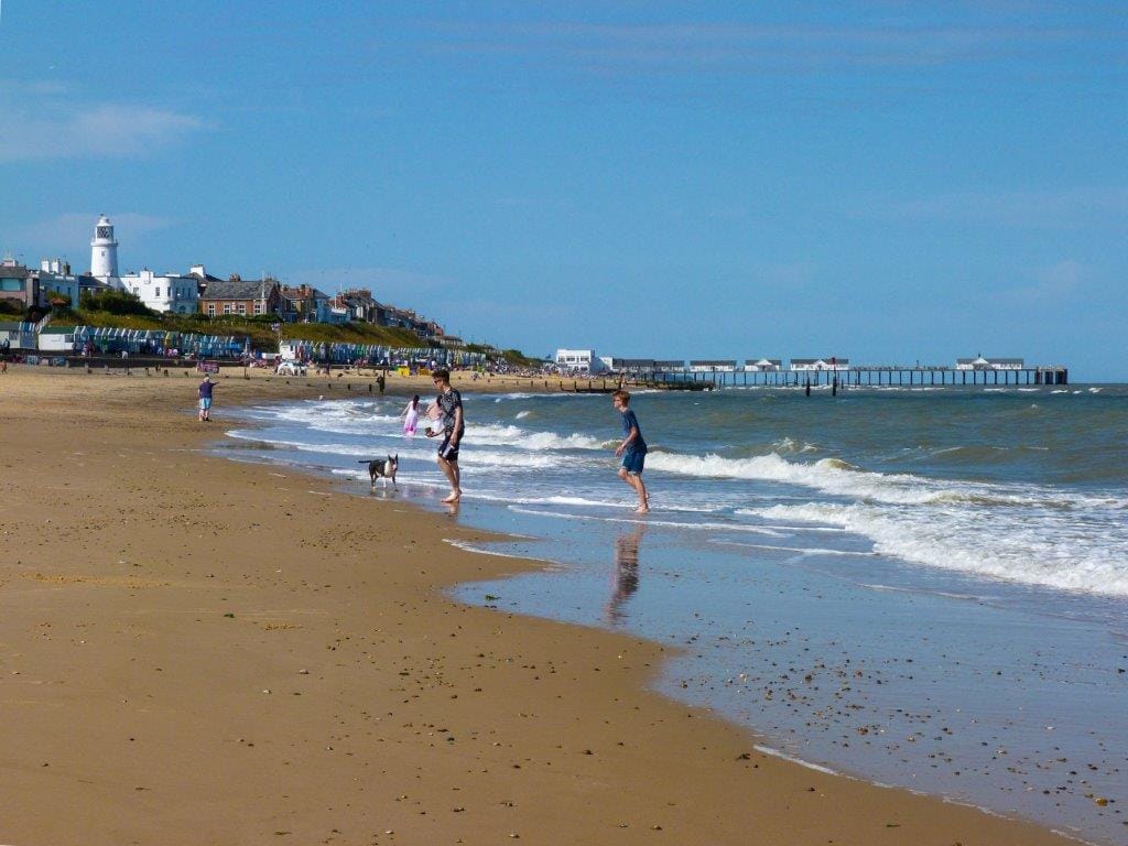 A sandy beach is depicted with gentle waves lapping at the shore. People can be seen enjoying the water, with a lighthouse and pier in the background, contributing to the coastal ambiance. The sky is bright with a few clouds, enhancing the beach's inviting atmosphere.