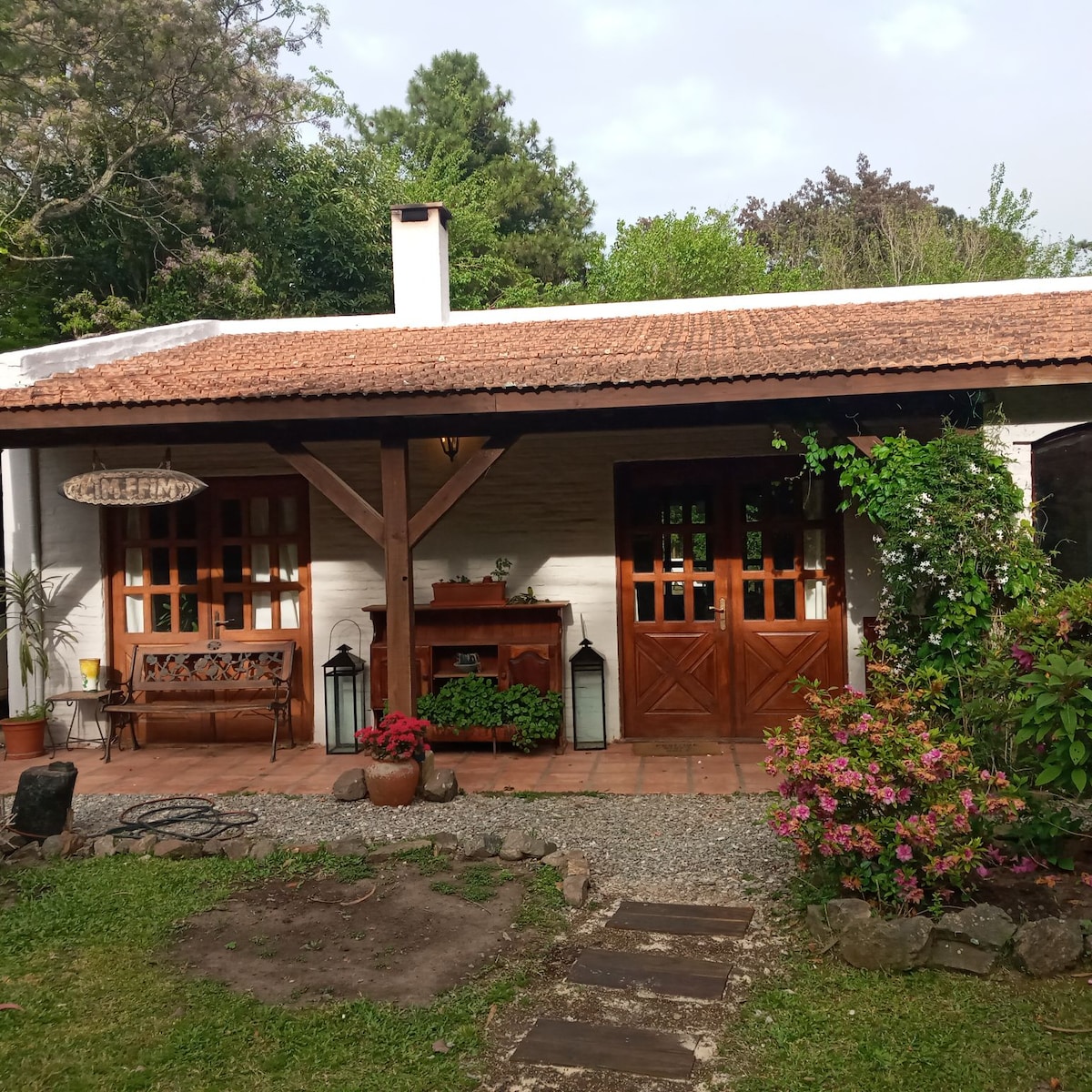 The exterior entrance features a wooden porch with a bench, surrounded by colorful flowers. Two sets of double doors open into the home, complemented by decorative lanterns and potted plants. The natural greenery creates a serene and inviting ambiance.