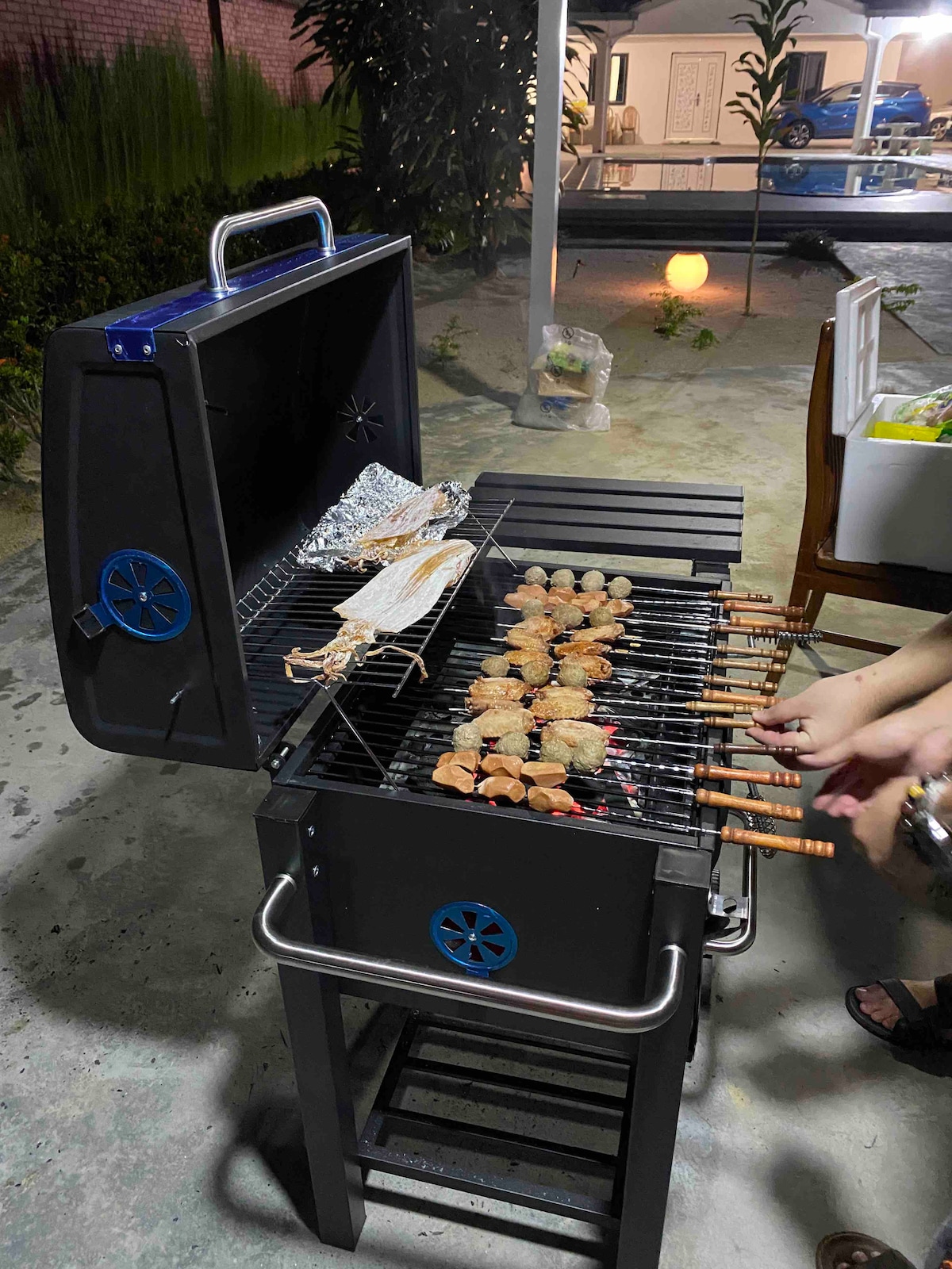 A black grill is positioned outdoors, displaying various skewered meats being grilled. The grill lid is raised, and foil-wrapped items are visible on one side. In the background, greenery and additional outdoor elements can be seen, creating a relaxed atmosphere.