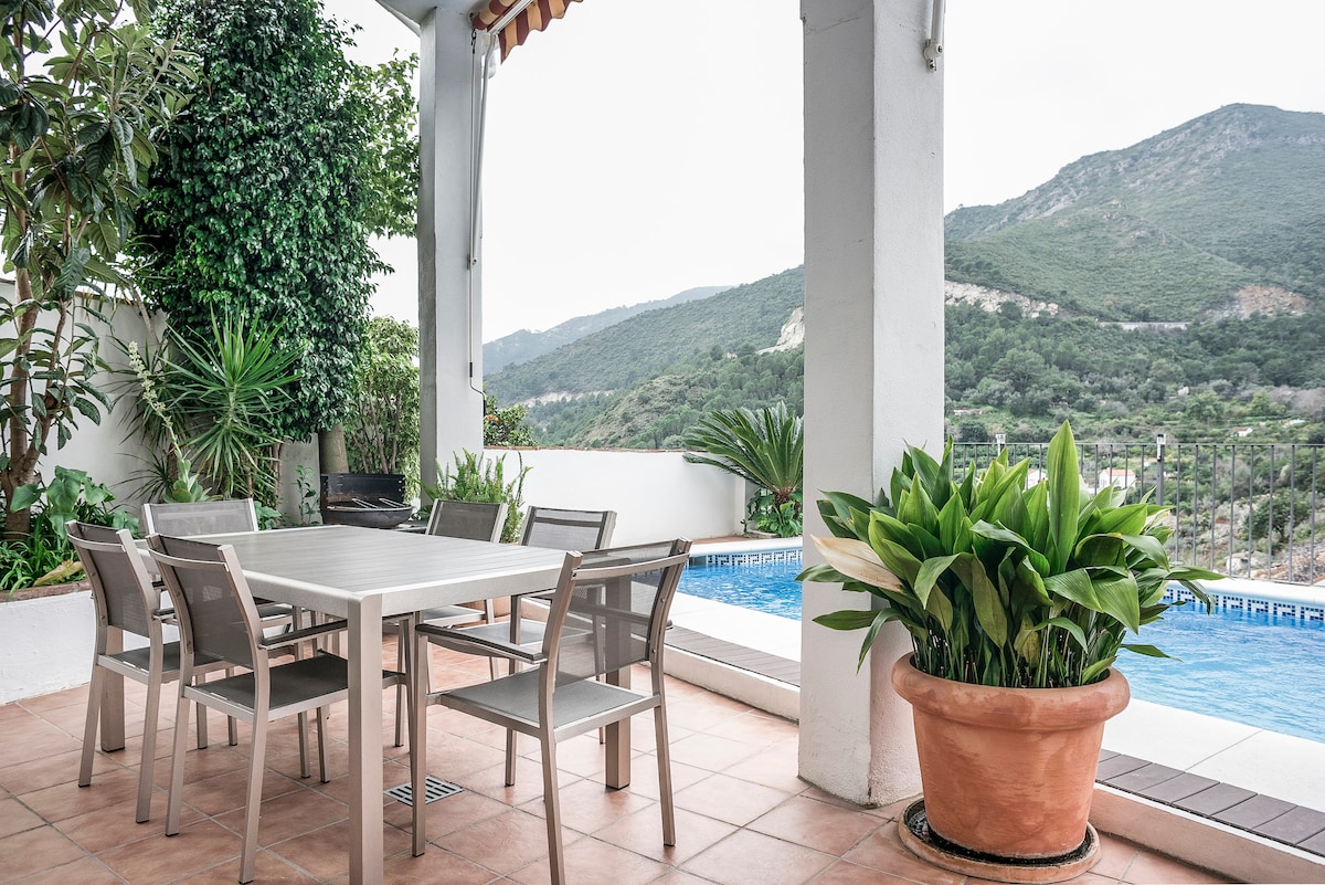 A covered terrace features a dining table surrounded by six chairs, inviting outdoor meals. Lush greenery and a large potted plant enhance the space, while views of the mountainous landscape and a private swimming pool are visible in the background.