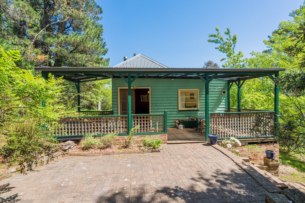 A charming weatherboard cottage is embraced by lush greenery, with a welcoming front verandah surrounded by trees. The spacious outdoor area features a paved entrance and offers a peaceful, private setting. A large window reflects a bright interior, enhancing the inviting character of the cottage.