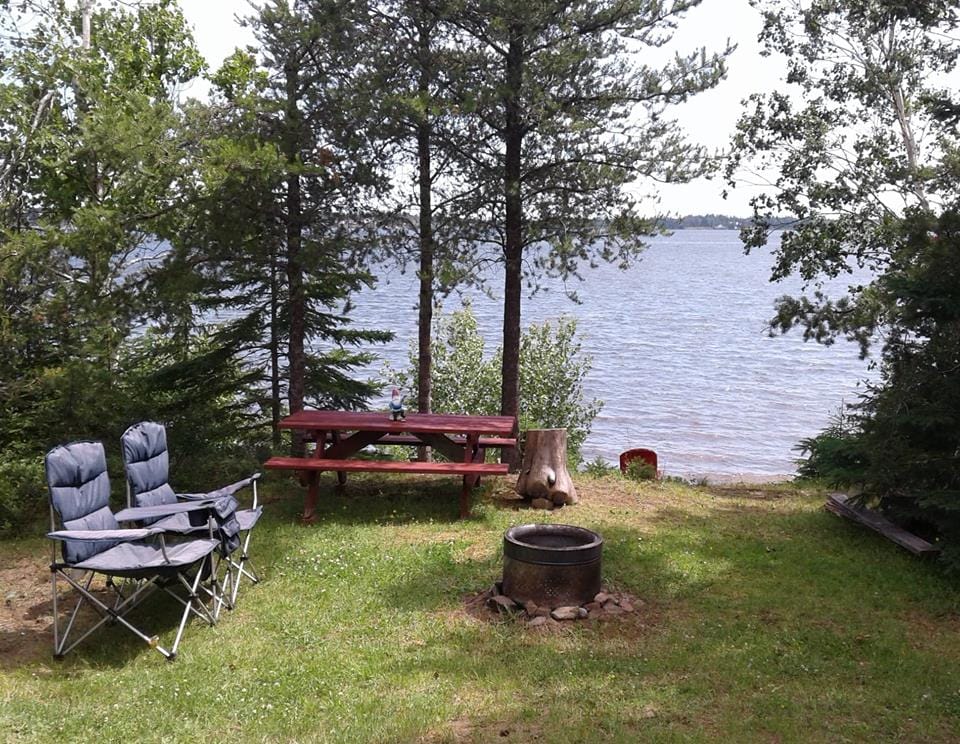A serene outdoor space by the water features a picnic table crafted from natural wood, accompanied by two comfortable folding chairs. A fire pit is arranged on a patch of grass, surrounded by trees, offering a peaceful view of the lake beyond.