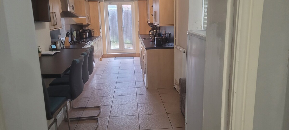 A well-equipped kitchen is highlighted with wooden cabinetry and dark countertops. The space features a breakfast bar with seating for several guests. Large glass doors provide access to a backyard, allowing natural light to fill the room, and tiled flooring extends throughout the area.