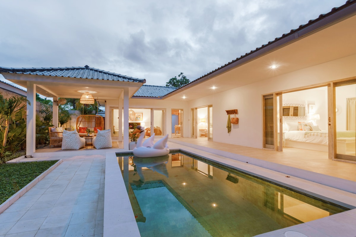 An inviting outdoor area features a private pool reflecting the sky, surrounded by tiled flooring and lush greenery. Adjacent, open living spaces showcase cozy seating and light-colored decor, with large glass doors allowing for maximum natural light and flow between indoor and outdoor environments.