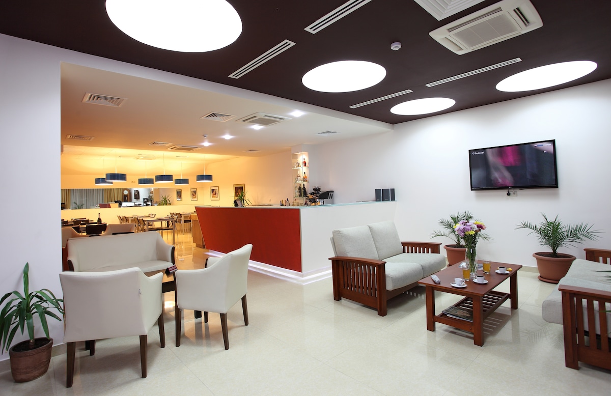 A modern lounge area is presented with neutral furniture, featuring a wooden table and comfortable seating. The space is illuminated by circular ceiling lights, enhancing the clean lines of the design. Potted plants add greenery, while a small bar area and a television are visible in the background.