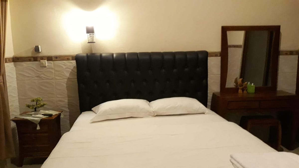 A cozy sleeping area features a soft bed with a tufted black headboard. Natural light is provided by a wall lamp above the bed, while a small wooden nightstand and a mirror with a light wooden frame are positioned nearby. White linens complement the neutral-toned walls.