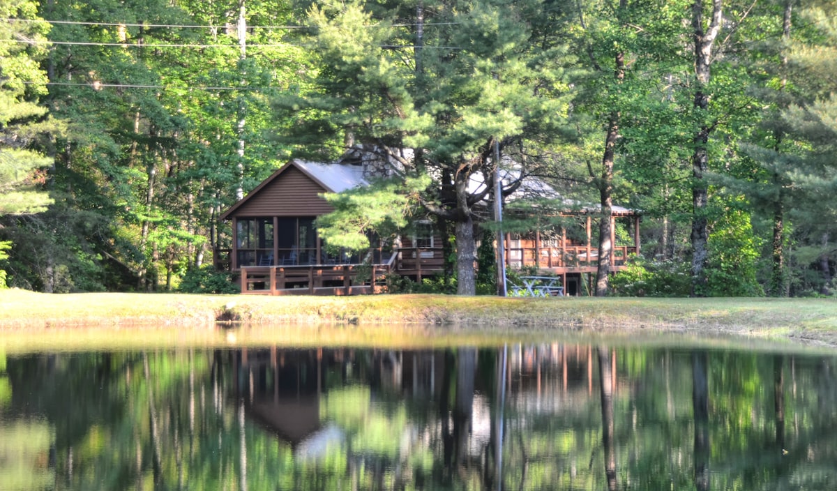 A log cabin is nestled among tall trees, showcasing a deck that overlooks a serene pond. The tranquil water reflects the cabin and surrounding greenery, creating a peaceful natural setting. Outdoor seating is visible on the deck, inviting relaxation by the water.