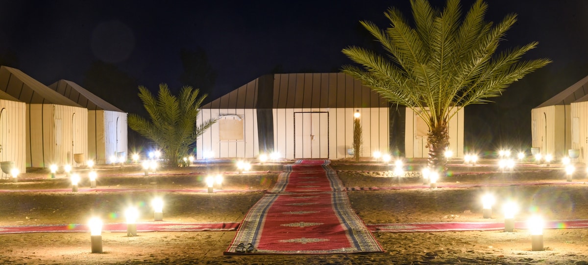 A pathway illuminated by soft lights leads to the white tents, set amidst palm trees. The spacious surroundings are decorated with lanterns, creating a welcoming ambiance under the night sky. The tents feature individually lockable doors, enhancing privacy and security.
