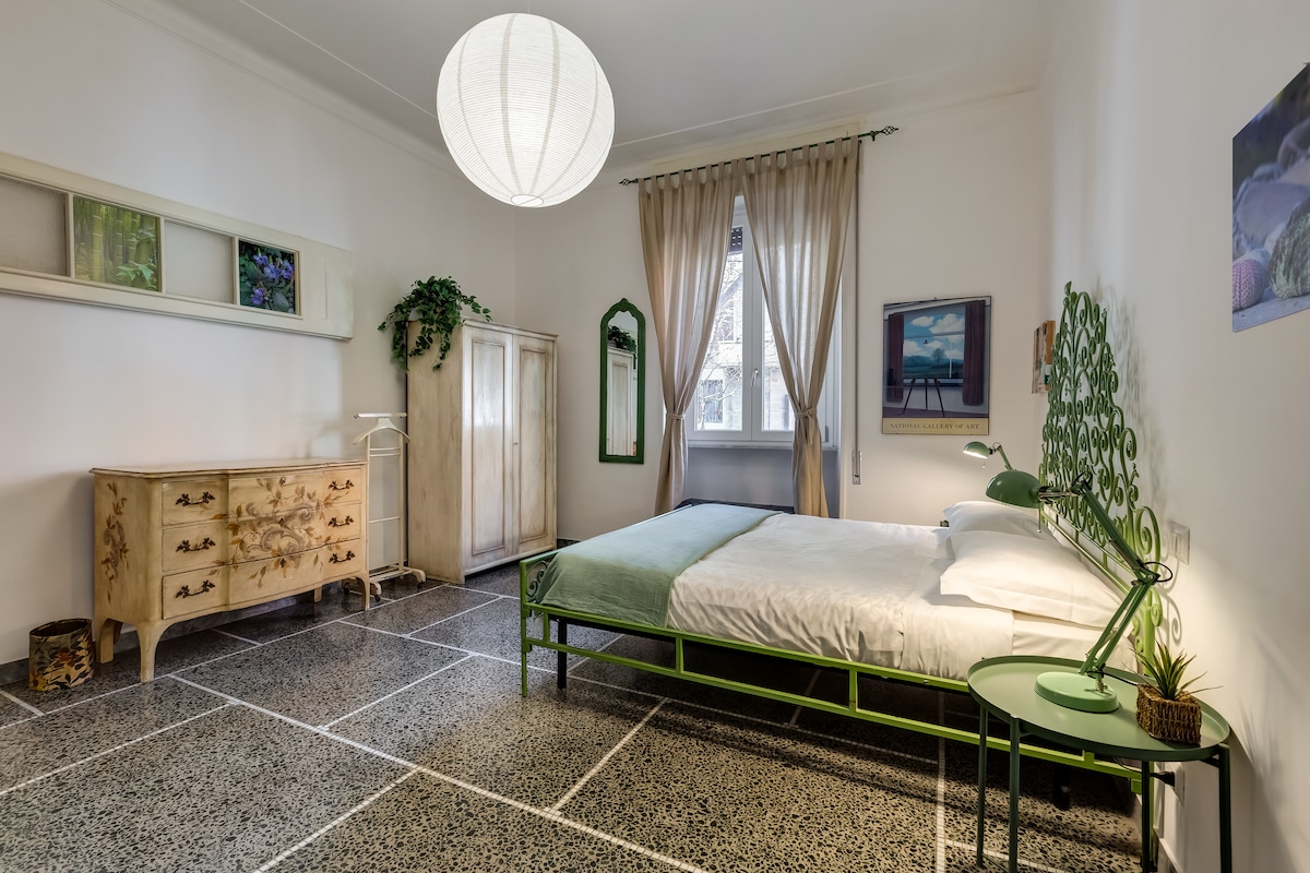 A spacious bedroom features a King size bed with a light green bedspread. An antique dresser and a wardrobe stand against the wall. Natural light enters through a large window, with soft drapes. A unique green headboard and a matching side table add color to the space.