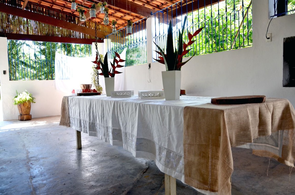 An open-air dining area features a long table covered with a light tablecloth. Potted plants with green foliage and red leaves are placed at either end, while large windows allow natural light to fill the space. The concrete floor and rustic ceiling complement the tranquil setting.