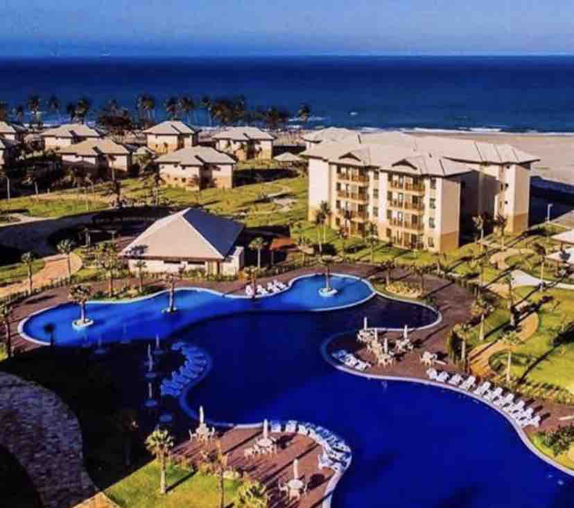 An expansive view of a resort complex is presented, featuring a large, winding pool surrounded by well-maintained gardens. Beachfront access is visible alongside a serene ocean in the background, with palm trees adding a natural touch to the landscape.