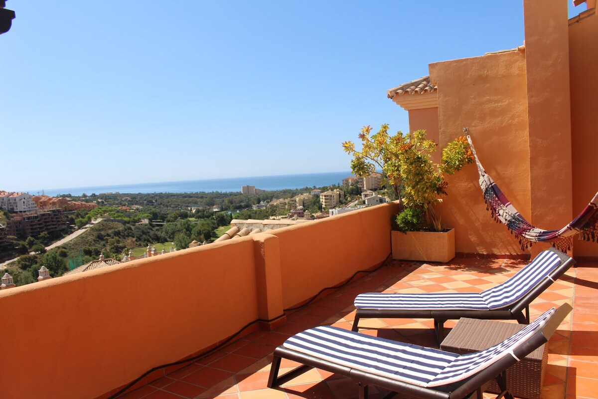 An expansive terrace features two striped sun loungers and a colorful hammock. A potted plant adds a touch of greenery. The view encompasses the coastline and surrounding hills, under clear blue skies, providing a tranquil setting for relaxation.