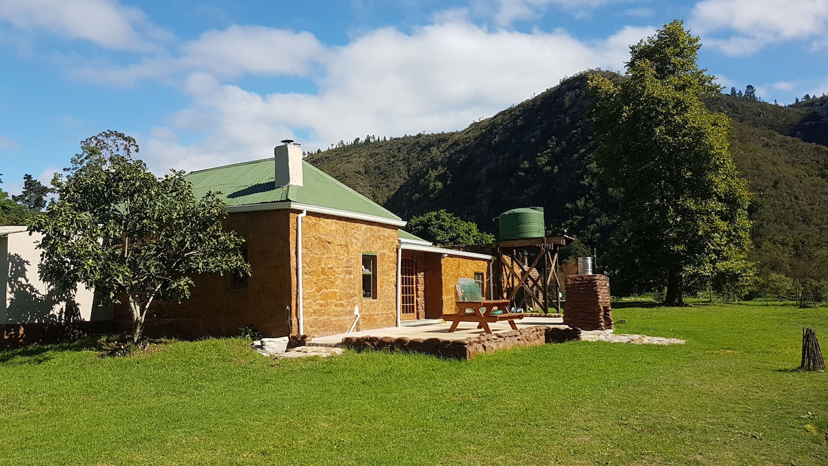 The exterior of the cottage features a natural stone facade with a green roof surrounded by lush grass. A water tank is positioned near the building, and a wooden picnic table is located on the terrace, providing a peaceful outdoor space. The backdrop includes rolling hills and greenery.