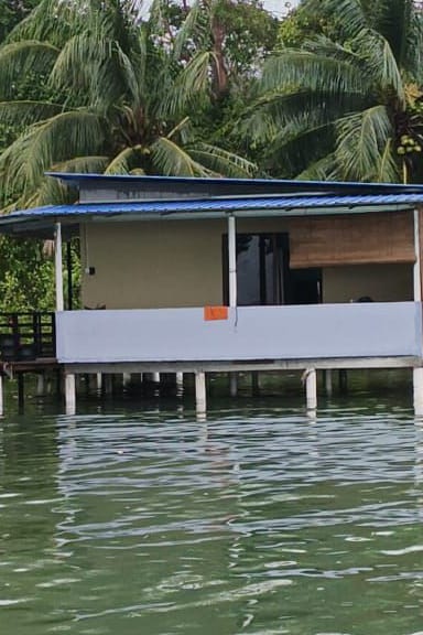 A tranquil waterfront property is depicted, supported by stilts above the calm water. The exterior features a blue roof and light-colored walls, with large windows offering views of the surrounding nature, including lush palm trees in the backdrop.