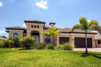The property features a modern architectural design with a tiled roof and decorative archways. Lush green grass and tropical plants surround the entrance, leading up to a spacious driveway suitable for multiple vehicles.