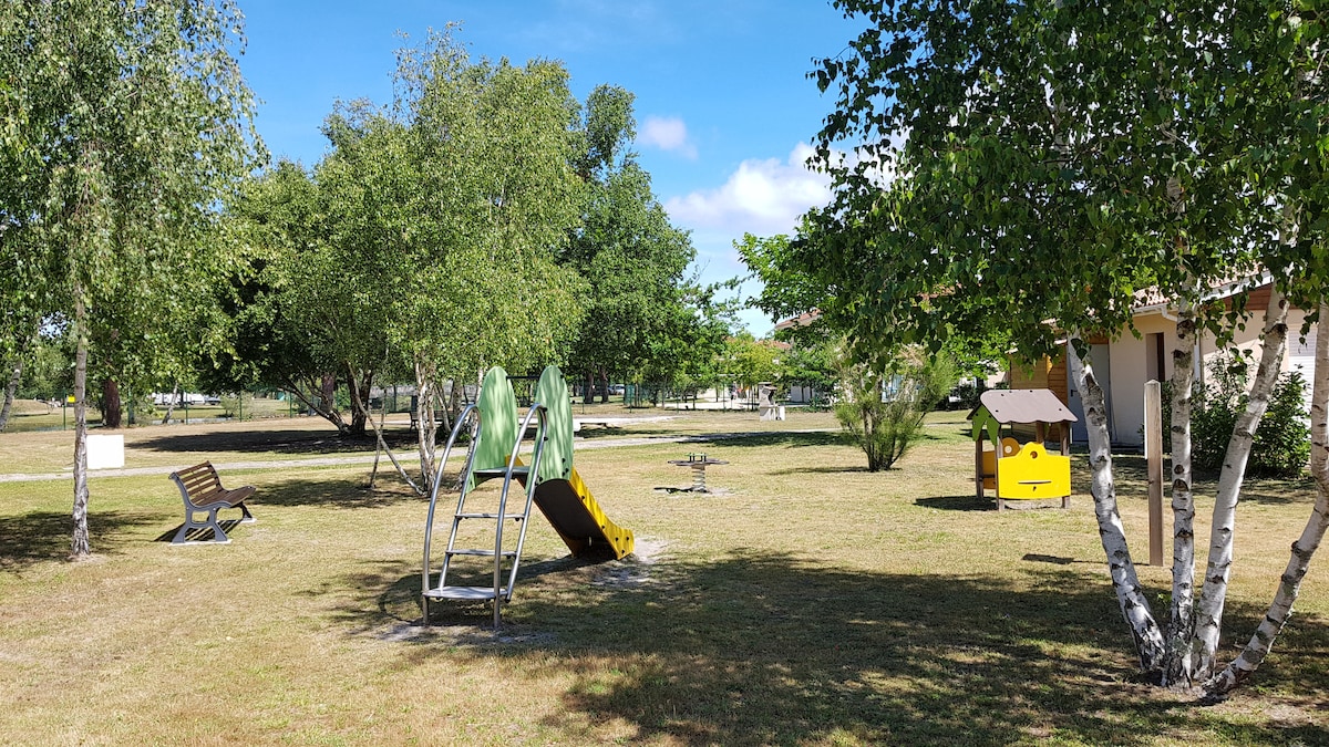 A spacious grassy area features a playground with a slide and climbing frame, surrounded by trees. A bench is positioned nearby, offering a place to sit and relax. A playhouse and a small structure for games are also visible in the background.