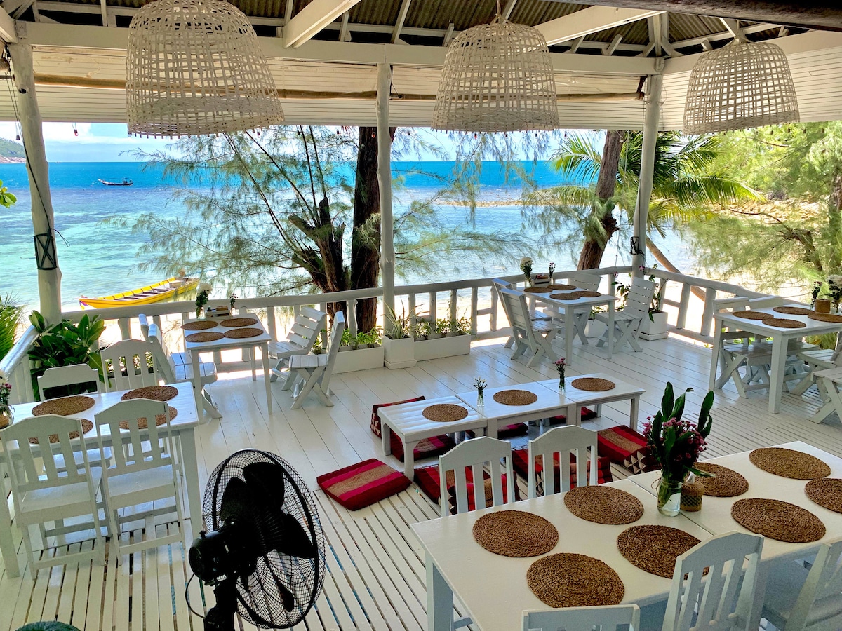 A spacious beachfront dining area features white wooden tables and chairs, arranged around woven placemats. Natural light fills the space through open walls, with views of the calm blue sea and nearby palm trees. Decorative pendant lights hang from the ceiling, adding to the inviting atmosphere.