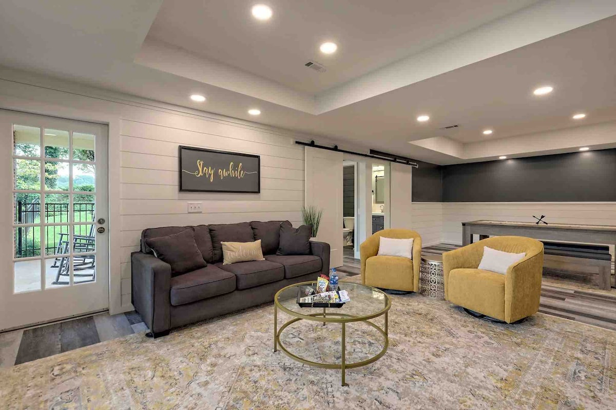 A spacious living area features a comfortable dark sofa and two yellow armchairs. Natural light enters through the glass doors, which lead to an outdoor patio. A round coffee table sits in the center, adorned with small decor items, on a patterned area rug.