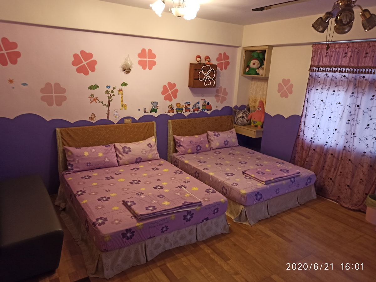 A bright sleeping area features two spacious beds adorned with floral-patterned linens. Pink and purple accents decorate the walls, complemented by playful artwork and a window with sheer curtains allowing natural light. A small seating area is present, enhancing the welcoming environment.