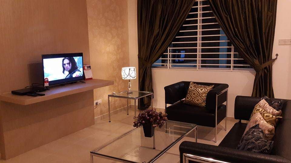A modern living area features two sleek black couches adorned with decorative pillows. A glass coffee table occupies the center, while a small side table with a lamp adds illumination. A television is mounted on the wall, and soft light filters through the window covered with dark curtains.