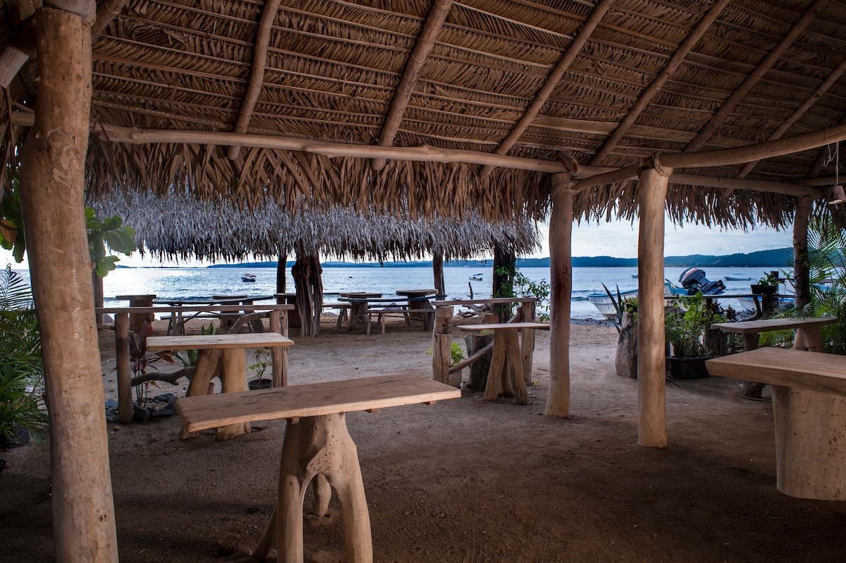An open-air palapa is visible, featuring several wooden tables surrounded by natural seating made from tree trunks. The sandy floor leads towards the water's edge, where distant boats are anchored, and the tranquil bay is seen in the background under a soft evening light.