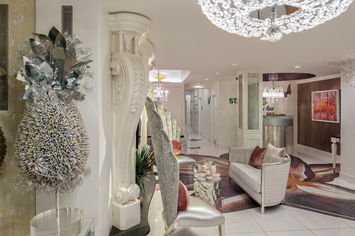 The lobby features modern seating arrangements with plush armchairs and a decorative rug. Unique art pieces, including a large ornamental sculpture, add character to the space. A shimmering chandelier provides illumination, while the reception area is marked by a contemporary design and welcoming atmosphere.