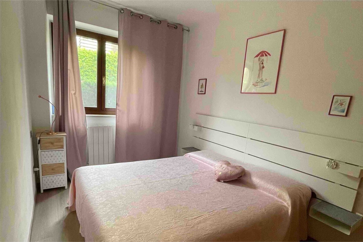 A softly lit bedroom features a spacious bed with a light pink coverlet. Pale pink curtains frame a window that offers a view of greenery outside. Wall art and a bedside table with storage complete the calming atmosphere of the room.
