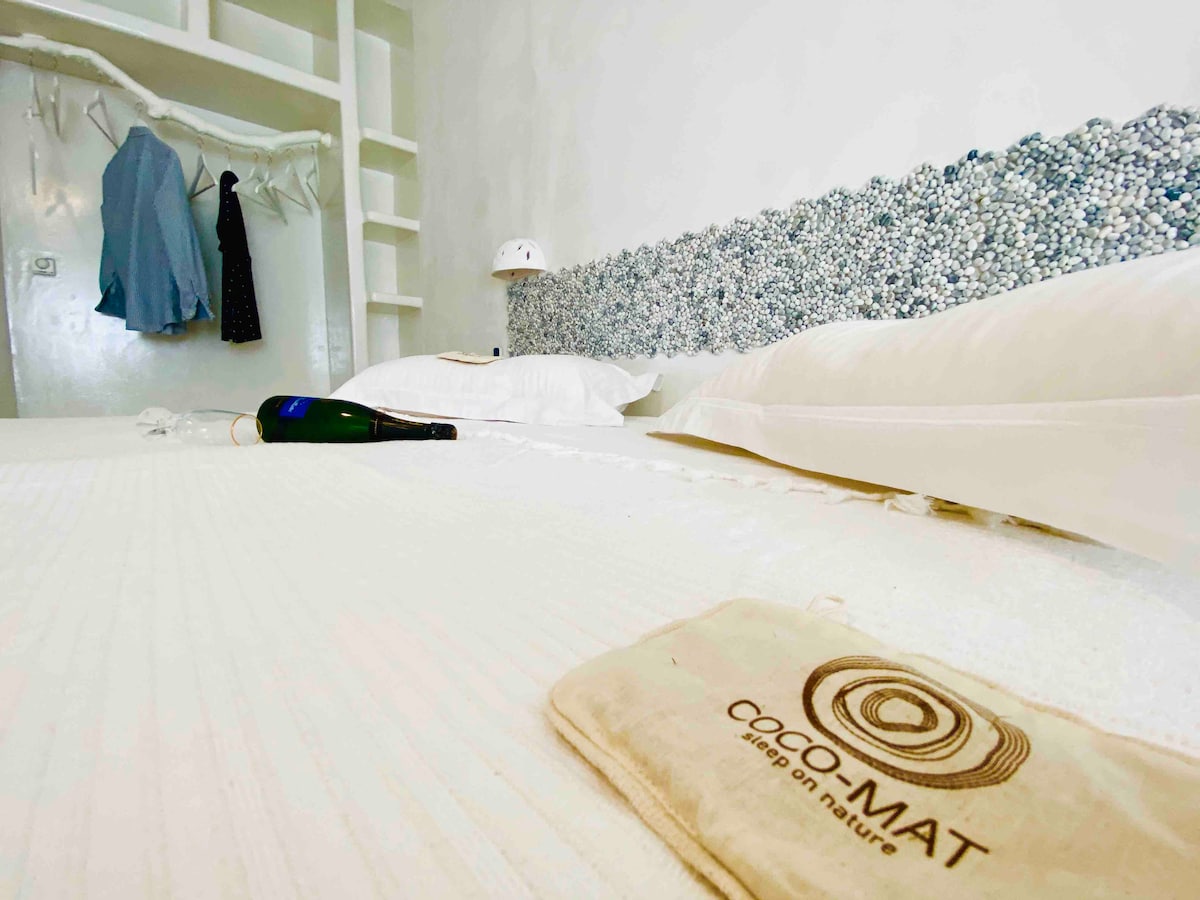 A neatly arranged sleeping area features a spacious bed with crisp white linens. Decorative elements include a textured headboard composed of small stones. Nearby, a bottle of champagne rests on the bed, and light-colored garments hang on a rack, adding a touch of elegance to the setting.
