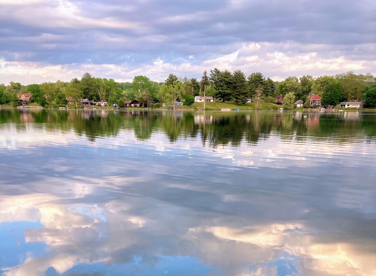 The tranquil surface of the spring-fed lake reflects lush green trees and a serene sky. Various cottages are visible along the shore, creating a peaceful lakefront scene surrounded by nature.