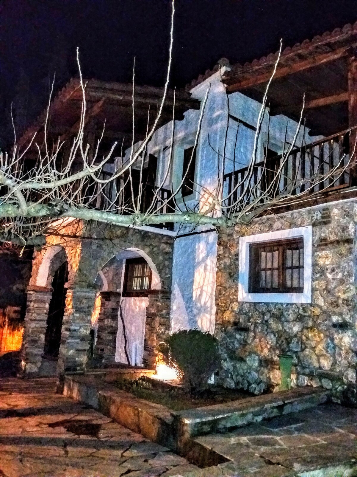 The exterior of a stone house is showcased at night, with its distinctive white walls illuminated. A wooden balcony extends from the upper level, and a stone pathway leads to the entrance. Leafless trees frame the scene, enhancing the rustic charm of the architecture.